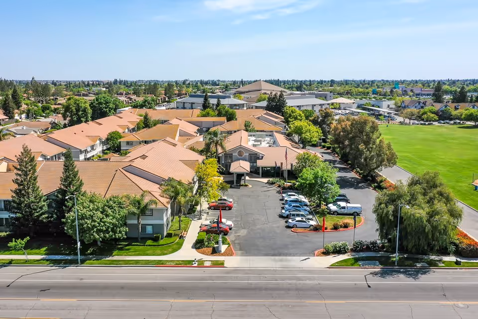 Aerial view of Fresno Senior Living facility showing multiple buildings with orange-tiled roofs, surrounded by trees and greenery. There is a parking lot with several cars in front of the main entrance, and a large green field adjacent to the property. The sky is clear and blue.