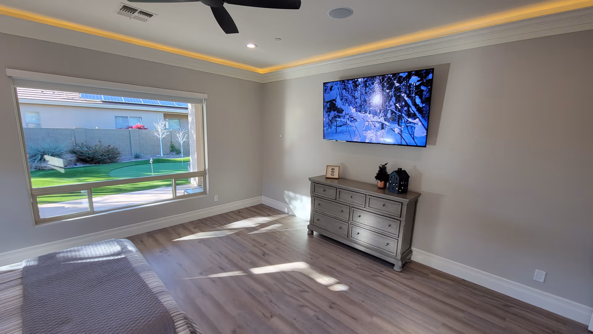 A bright and spacious living room with light wood flooring, a large window showing a green outdoor putting green, a wall-mounted flat screen TV displaying a snowy forest scene, and a gray dresser with decorative items on top.