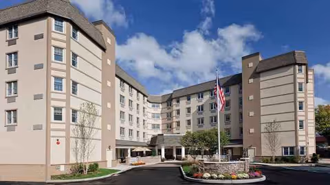 Atria Glen Cove exterior: a multi-story beige building with front entrance and flag.