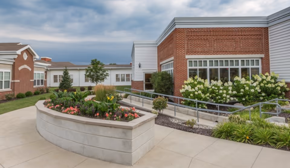 Exterior courtyard and entrance of a brick-and-siding retirement community building with landscaped flower beds and a wheelchair ramp.