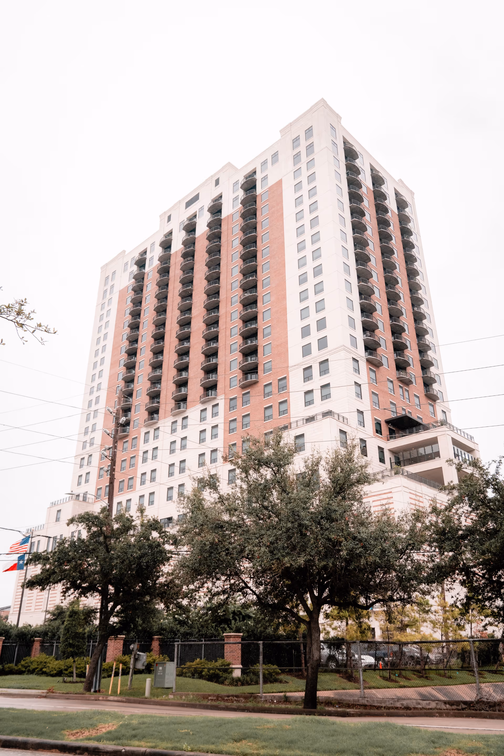 Tall multi-story residential building with balconies, surrounded by trees and a fenced area, under an overcast sky.