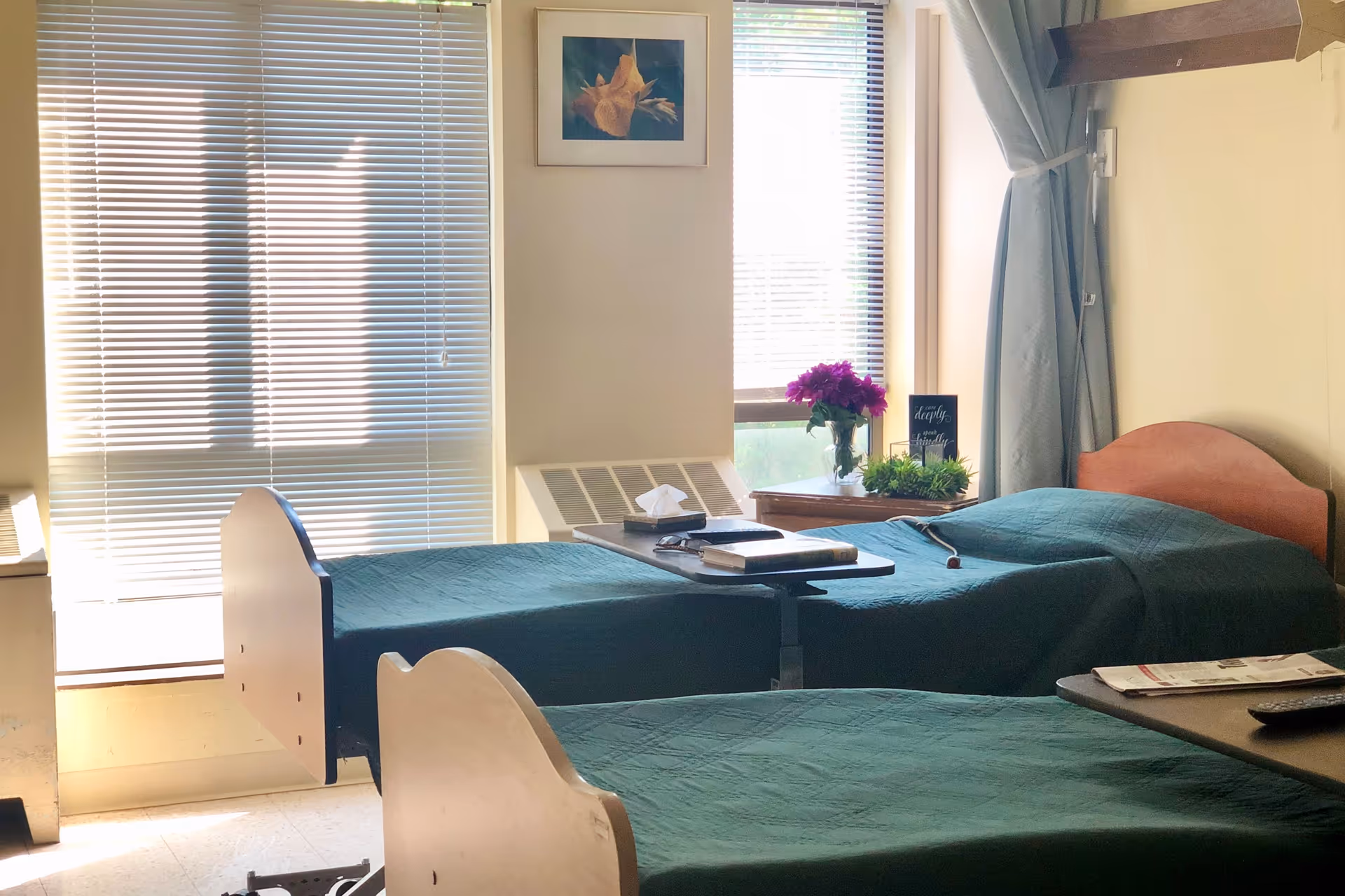 Two twin beds with green bedspreads in a bright patient bedroom with windows, tray tables, and a bedside table holding flowers.