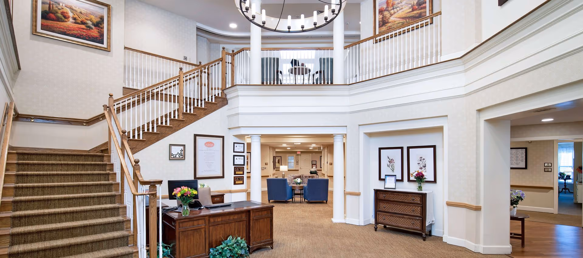 Bright two-story senior living lobby with a wooden staircase, reception desk, seating area, and framed artwork.