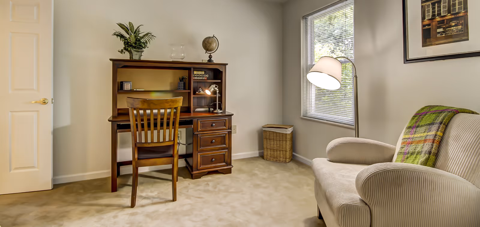A cozy room with a wooden desk and chair, a small table lamp, a plant, and a globe on the desk. There is a window with blinds letting in natural light, a wicker basket in the corner, and a cushioned armchair with a colorful plaid blanket draped over the back. A framed picture hangs on the wall above the armchair.