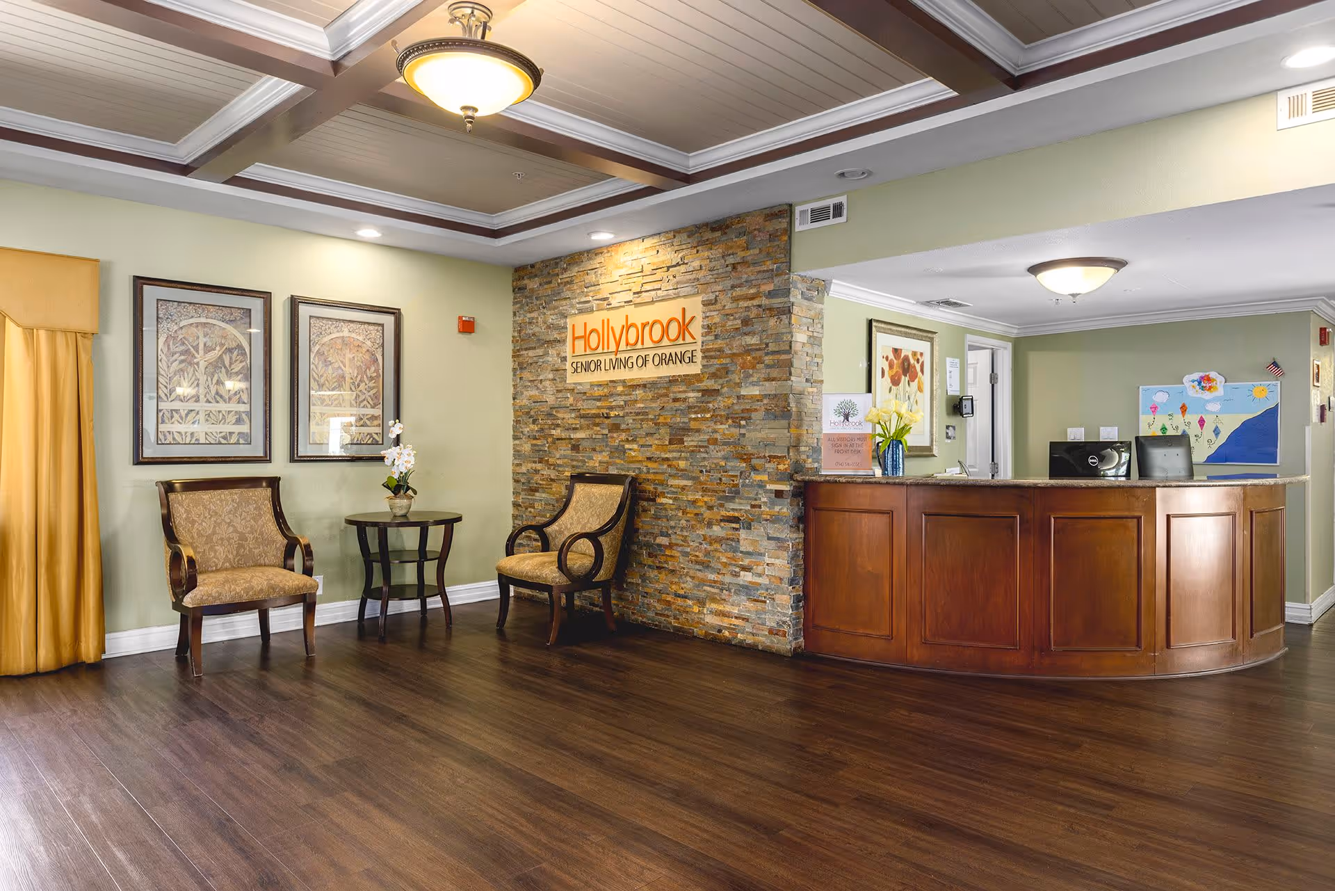 Reception area of a senior living facility with a wooden front desk, two upholstered chairs with a small round table between them, framed artwork on the walls, and a stone accent wall with a sign that reads 'Hollybrook Senior Living of Orange'.