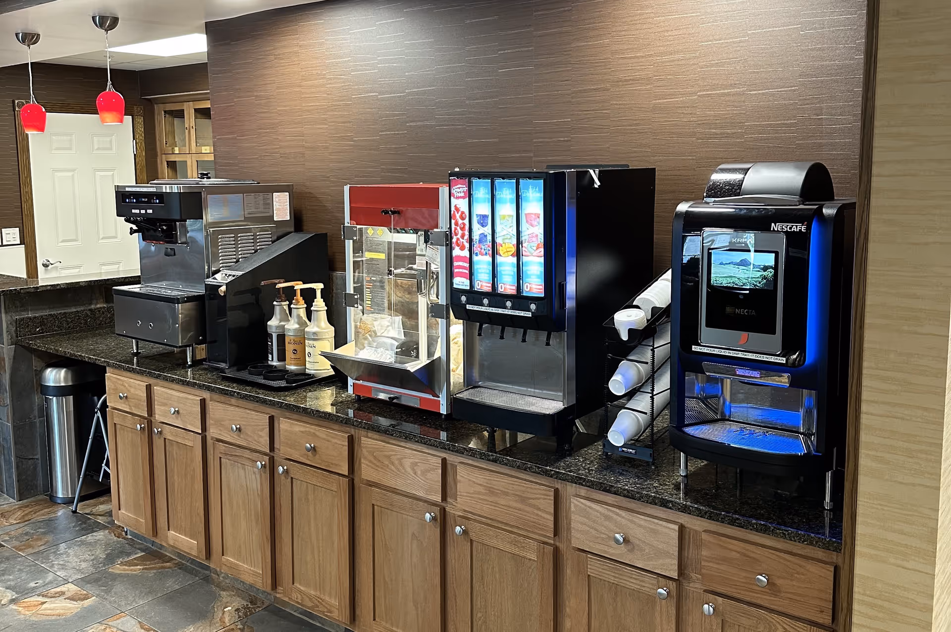 A countertop beverage station with multiple coffee machines, a soda dispenser, a popcorn maker, syrup bottles, and stacked disposable cups above wooden cabinets.