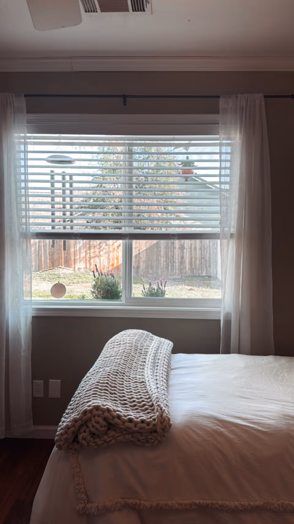 A neatly made bed with a folded chunky knit throw in front of a window with blinds and sheer curtains overlooking a backyard fence.