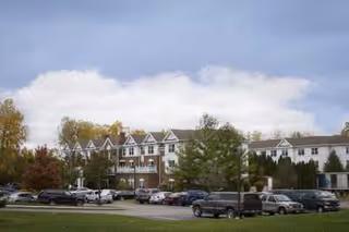 Exterior view of a multi-story senior living facility building with a parking lot in front, several cars parked, and trees surrounding the area under a partly cloudy sky.