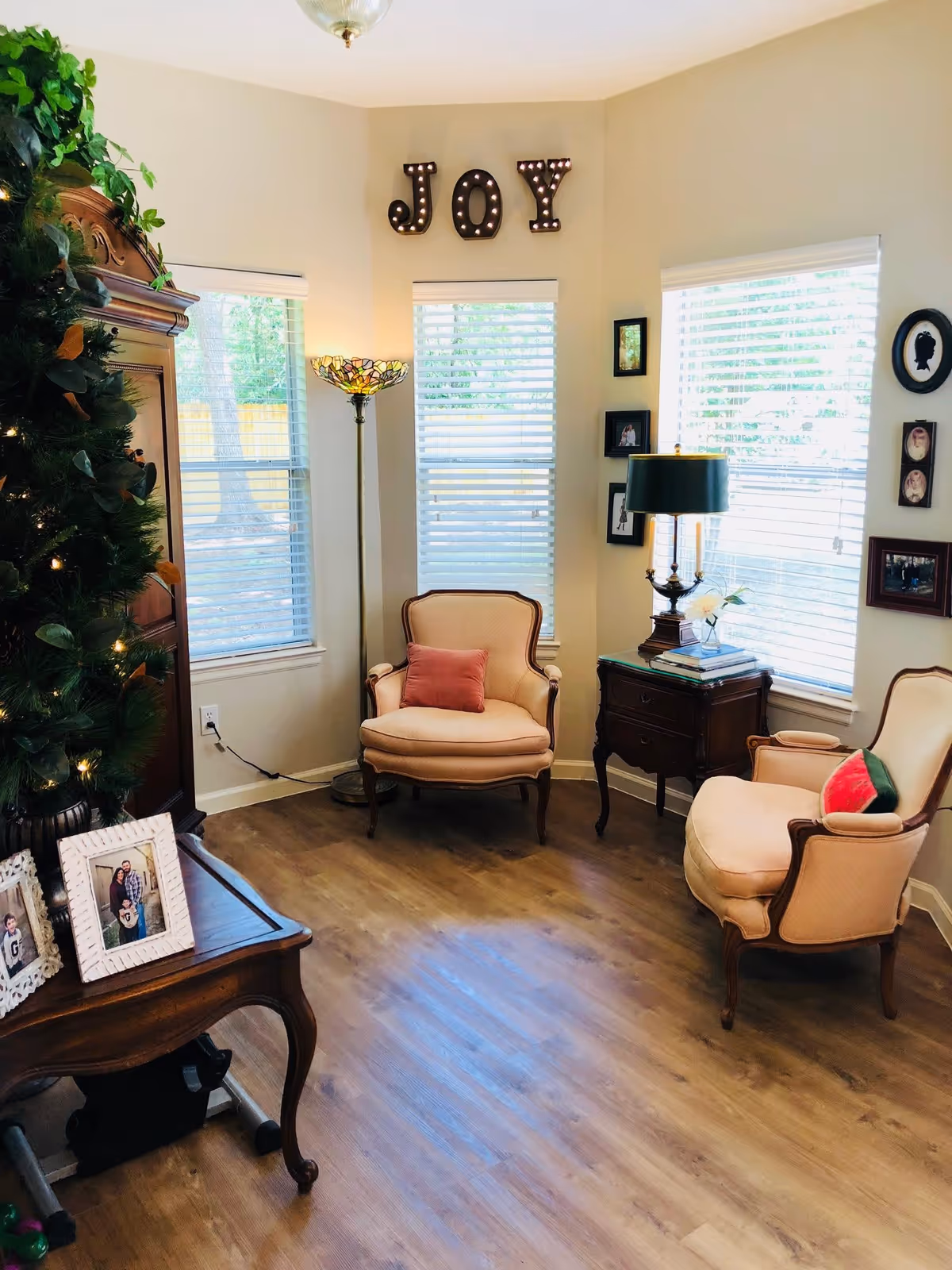A cozy living room corner with two beige upholstered armchairs, each with a decorative pillow. Between the chairs is a wooden side table with a green-shaded lamp and some books. Behind the chairs are three windows with white blinds letting in natural light. Above the middle window, the word 'JOY' is displayed in large marquee-style letters. To the left, part of a wooden cabinet and a decorated Christmas tree are visible. Family photos and framed artwork adorn the walls and a wooden table in the foreground holds framed photographs.