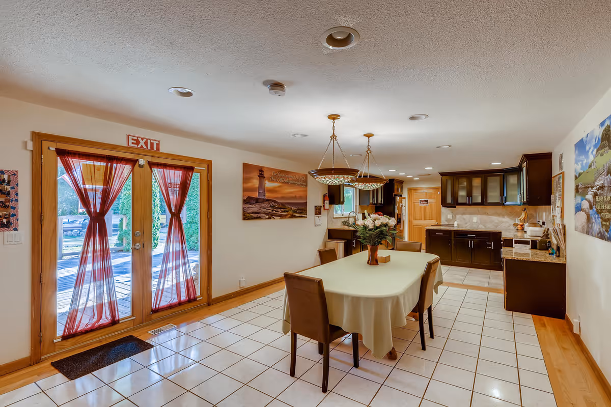 Interior view of a dining area and kitchen in a senior living facility. The dining area features a rectangular table covered with a light-colored tablecloth and surrounded by four chairs. A vase with flowers is placed on the table. Two pendant lights hang above the table. The kitchen area has dark wood cabinets, a countertop with various kitchen items, and a tiled backsplash. Double glass doors with red curtains lead outside, and there is an exit sign above the doors. The floor is tiled in the dining area and transitions to wood flooring near the kitchen.