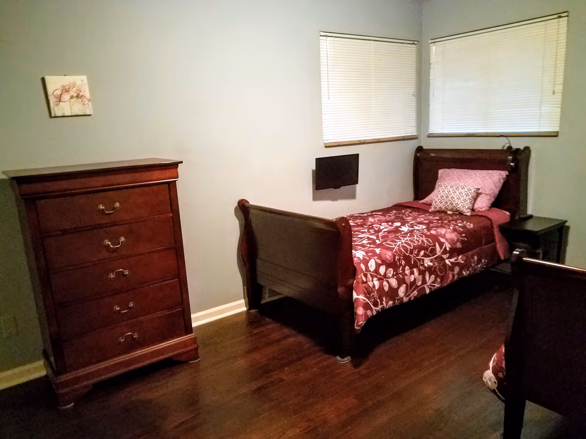 A small bedroom with a single wooden bed covered with a red floral bedspread and two pillows. Next to the bed is a dark wooden nightstand, and on the left side of the room is a tall wooden dresser with five drawers. Two windows with closed blinds are on the walls behind the bed. A small flat-screen TV is mounted on the wall near the bed. The floor is wooden.