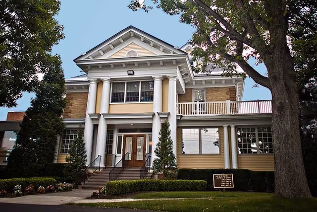 Front exterior view of a large, two-story senior living community building with classical architectural features including white columns, a triangular pediment, and a balcony. The building is surrounded by trees, shrubs, and a well-maintained lawn under a clear blue sky.