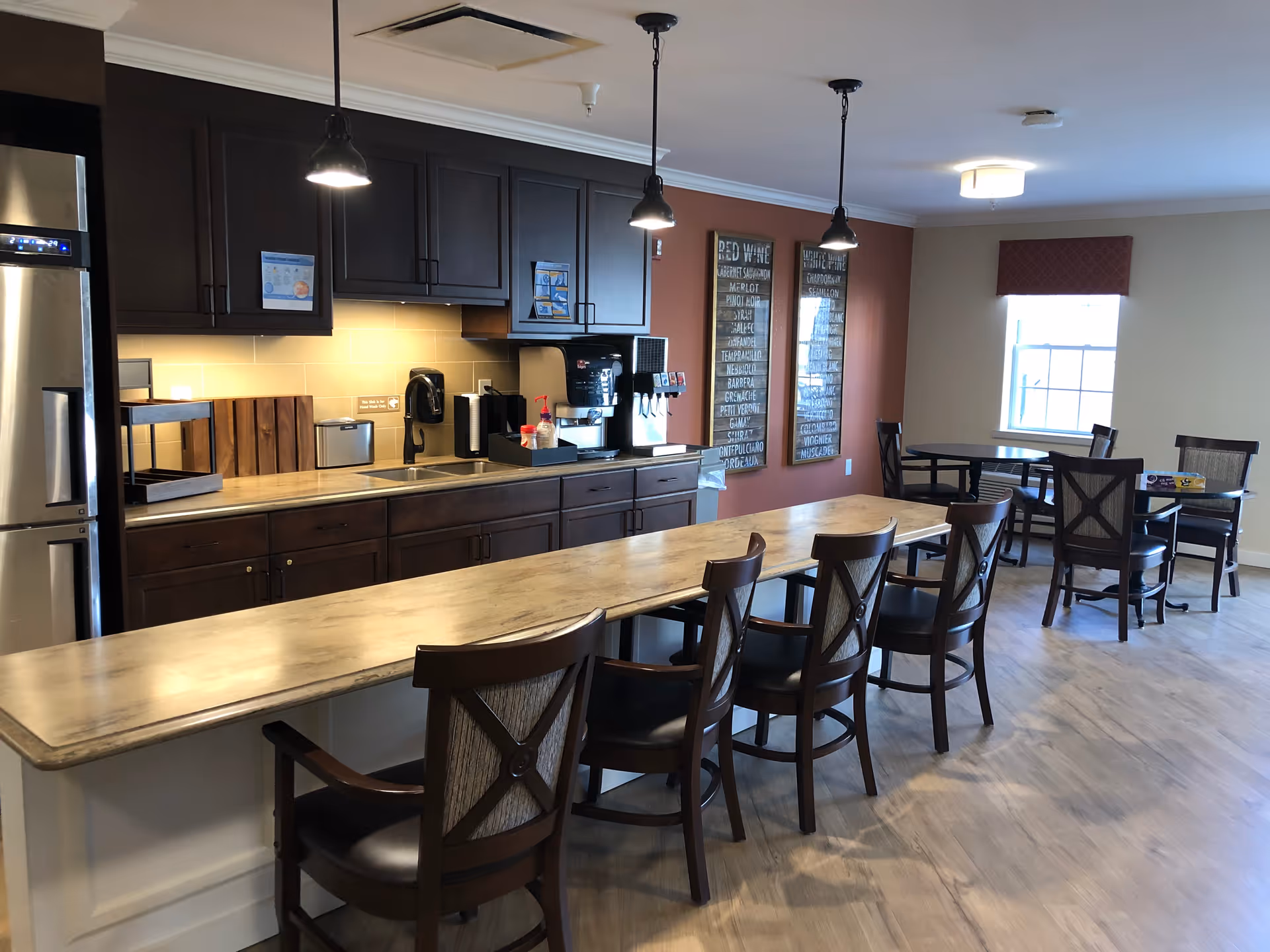 A senior living facility kitchen and dining area with a long counter and six wooden chairs. The kitchen features dark wood cabinets, a stainless steel refrigerator, a coffee machine, and under-cabinet lighting. Two framed wine lists hang on a red accent wall, and there are two round dining tables with chairs near a window with a red valance.