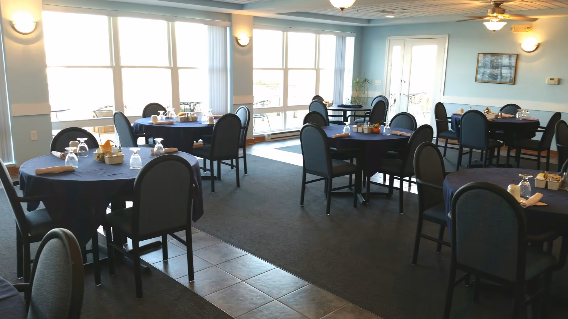 Dining room with multiple round tables covered with dark blue tablecloths, each set with upside-down glasses, napkins, and small condiment containers. The room has large windows letting in natural light, light blue walls, and ceiling fans with lights.