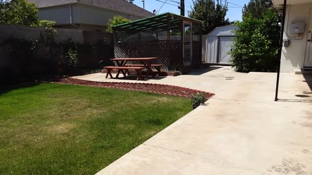 Outdoor area with a green lawn, a concrete pathway, a wooden picnic table with benches under a pergola, a small shed in the background, and some trees and bushes along the fence.