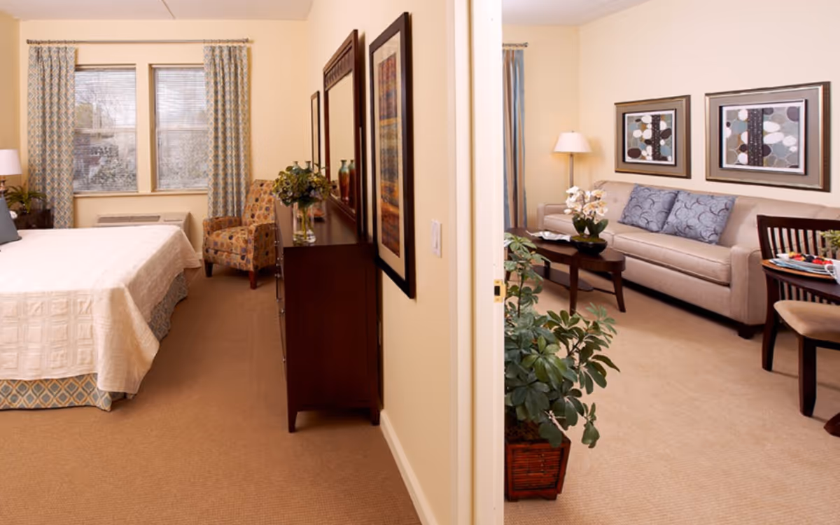 A cozy assisted living suite featuring a bedroom with a bed, patterned armchair, dresser with mirror, and window with curtains. Adjacent to the bedroom is a living room area with a beige sofa, coffee table with a flower arrangement, floor lamp, framed artwork on the walls, and a dining table with chairs. A potted plant is placed near the doorway between the two rooms.