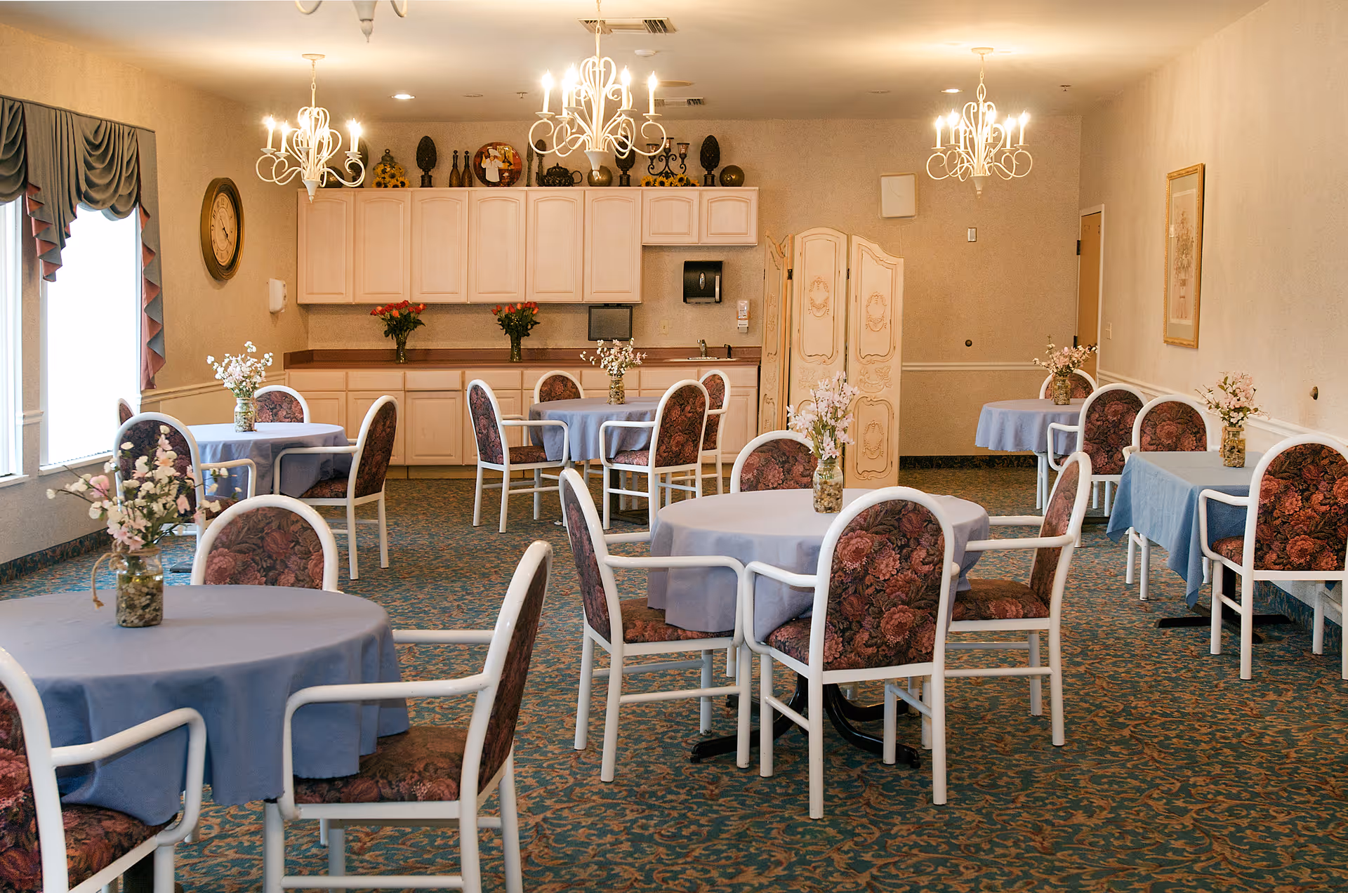 Dining room with round tables covered in blue tablecloths, floral centerpieces, upholstered chairs, chandeliers, and a serving counter in the back.
