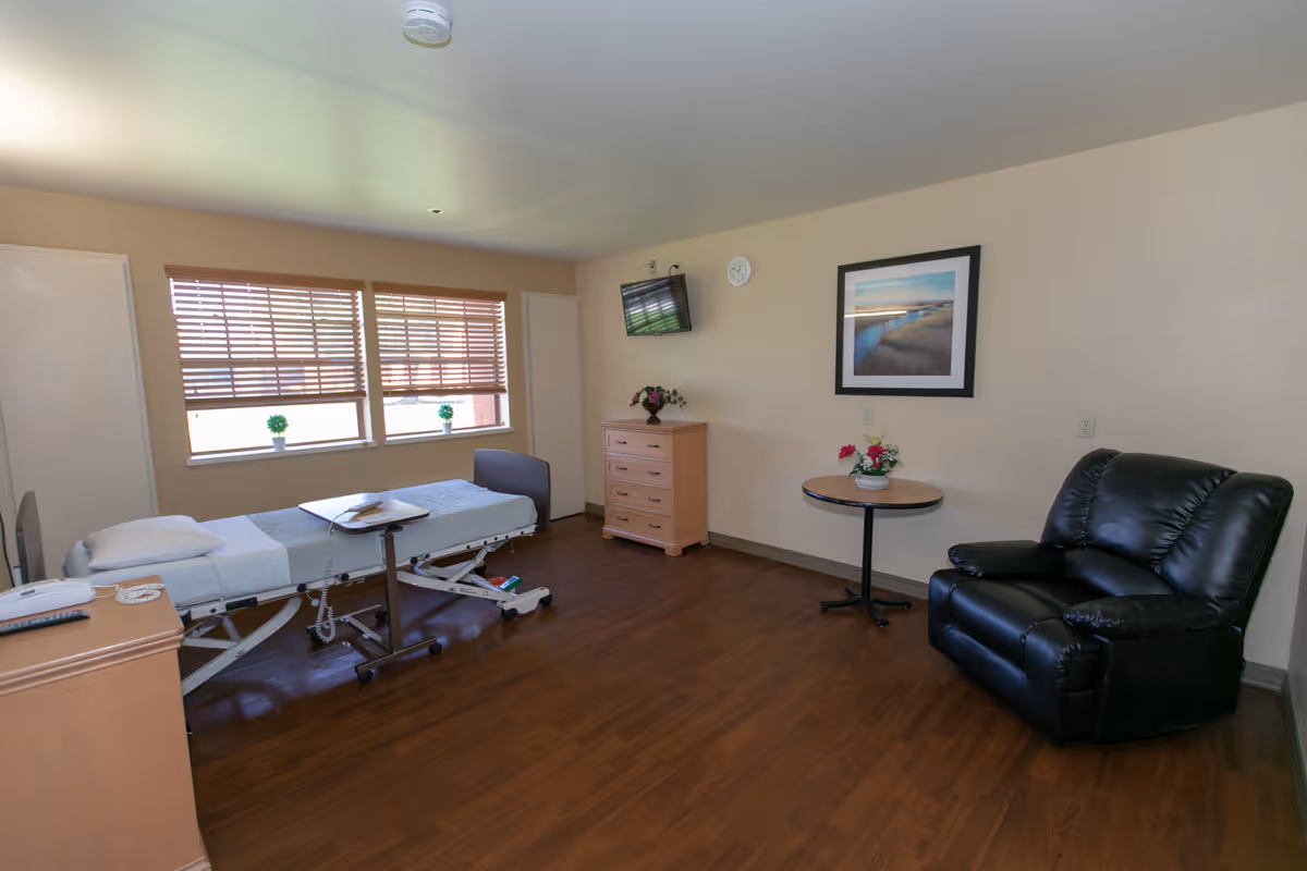 A room with a medical bed, a small wooden dresser, a round table with a flower vase, a black recliner chair, a wall-mounted TV, and two windows with blinds partially open letting in natural light. The floor is wooden and the walls are beige with a framed picture hanging.