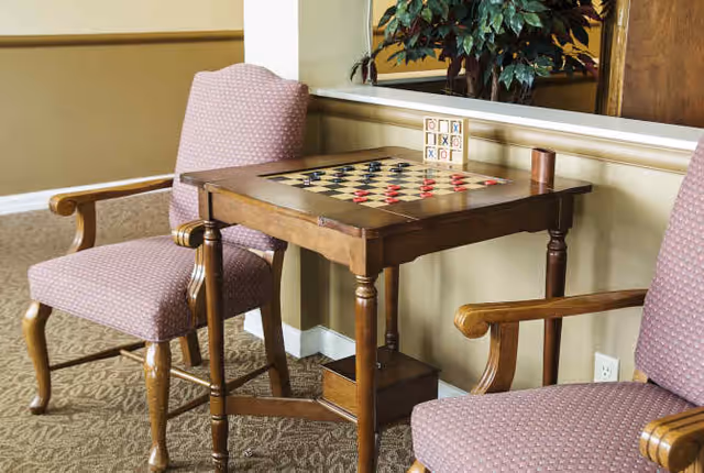 A wooden game table with a checkerboard and tic-tac-toe game set on top, flanked by two upholstered armchairs with wooden armrests, set against a beige wall with a large mirror and a potted plant in the background.