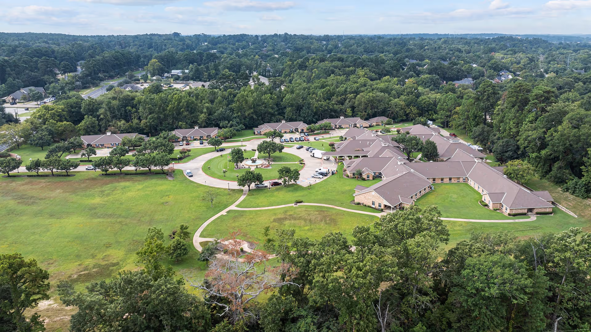 Aerial view of Atria Copeland senior living facility surrounded by green lawns and trees, with multiple single-story buildings arranged around a circular driveway with parked cars.