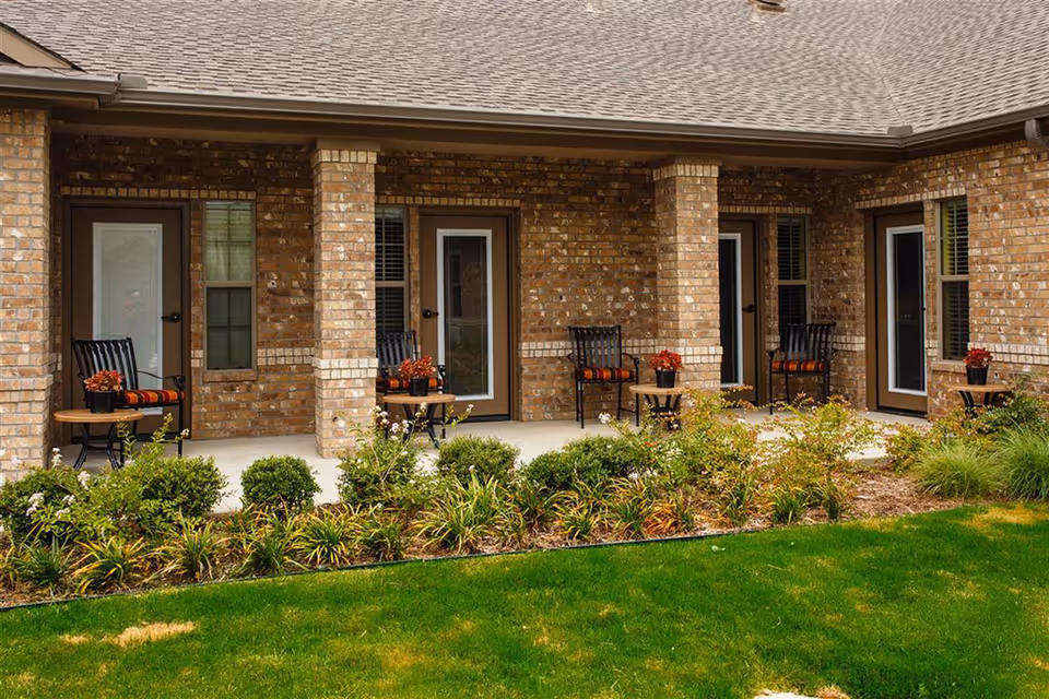 Outdoor patio area of a brick building with four black chairs, each paired with a small round table holding a potted plant with red flowers. The patio is bordered by a garden with green shrubs and plants, and a well-maintained lawn in the foreground.