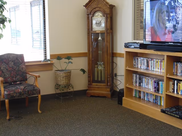 A cozy living room area with a floral upholstered armchair, a potted plant on a metal stand, a tall wooden grandfather clock, and a wooden entertainment center filled with DVDs and a television showing a woman on screen.
