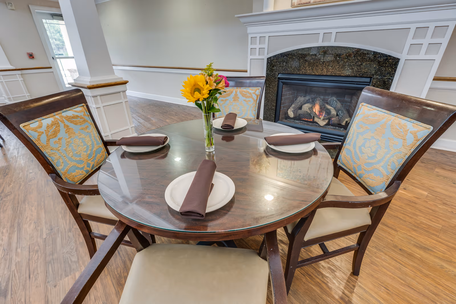A round wooden dining table with a glass top set with four plates and brown napkins, a vase with sunflowers and other flowers in the center, surrounded by four wooden chairs with patterned cushions. In the background, there is a lit fireplace with a white mantel and a wooden floor.