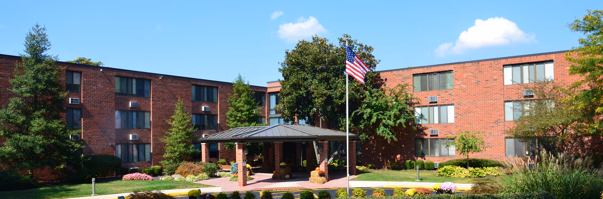 Exterior view of Brownsboro Park Retirement Community showing a three-story red brick building with multiple windows and air conditioning units. In front of the building is a covered entrance with a metal roof, surrounded by landscaped greenery, trees, and colorful flowers. An American flag is flying on a flagpole near the entrance under a clear blue sky.