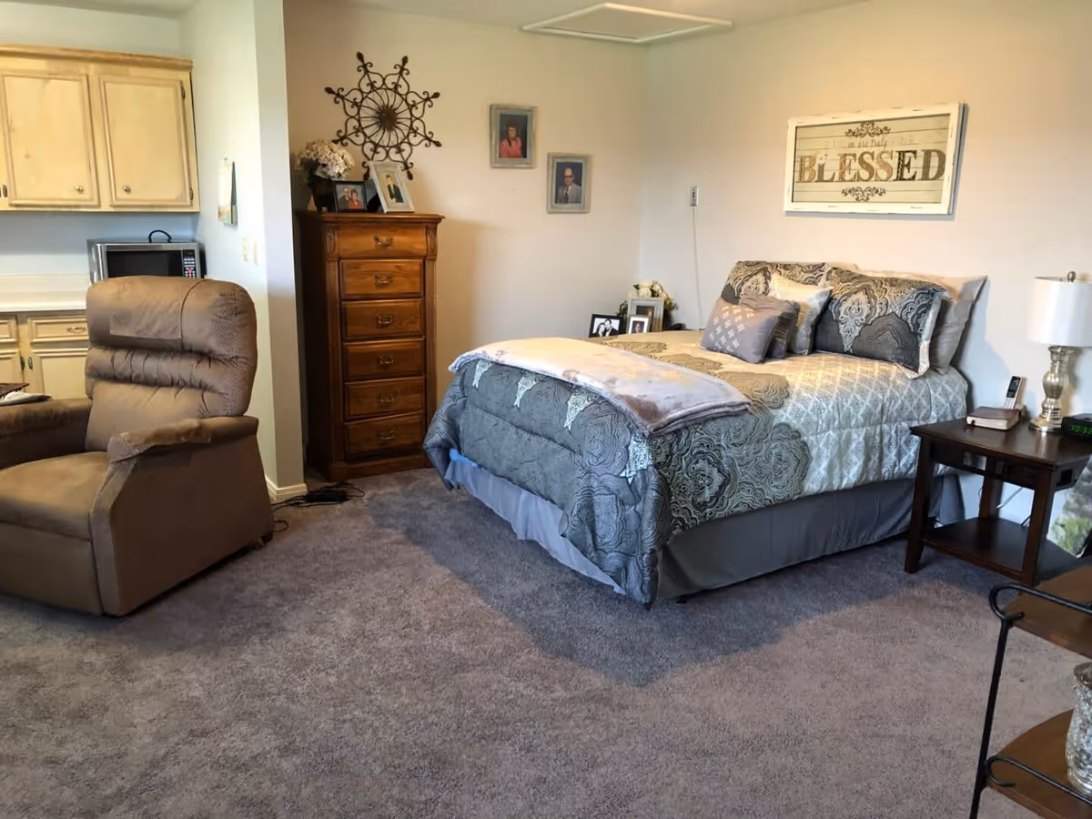 Furnished bedroom with a made bed and decorative pillows, a wooden dresser, nightstand with lamp, recliner, and a small kitchenette area.