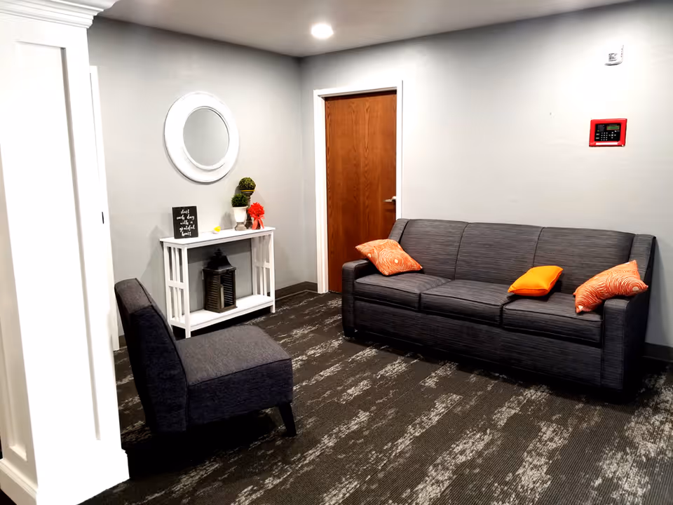 A small sitting area in a room with a gray couch adorned with three orange patterned pillows and one solid orange pillow, a dark gray upholstered chair, a white console table with decorative items including a round mirror above it, and a wooden door in the background.