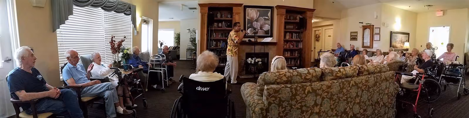 A group of elderly residents seated in a common living room area of a senior living facility, attentively listening to a man standing and speaking or singing near a fireplace with bookshelves on either side. The room is well-lit with large windows and decorated with floral artwork and plants.