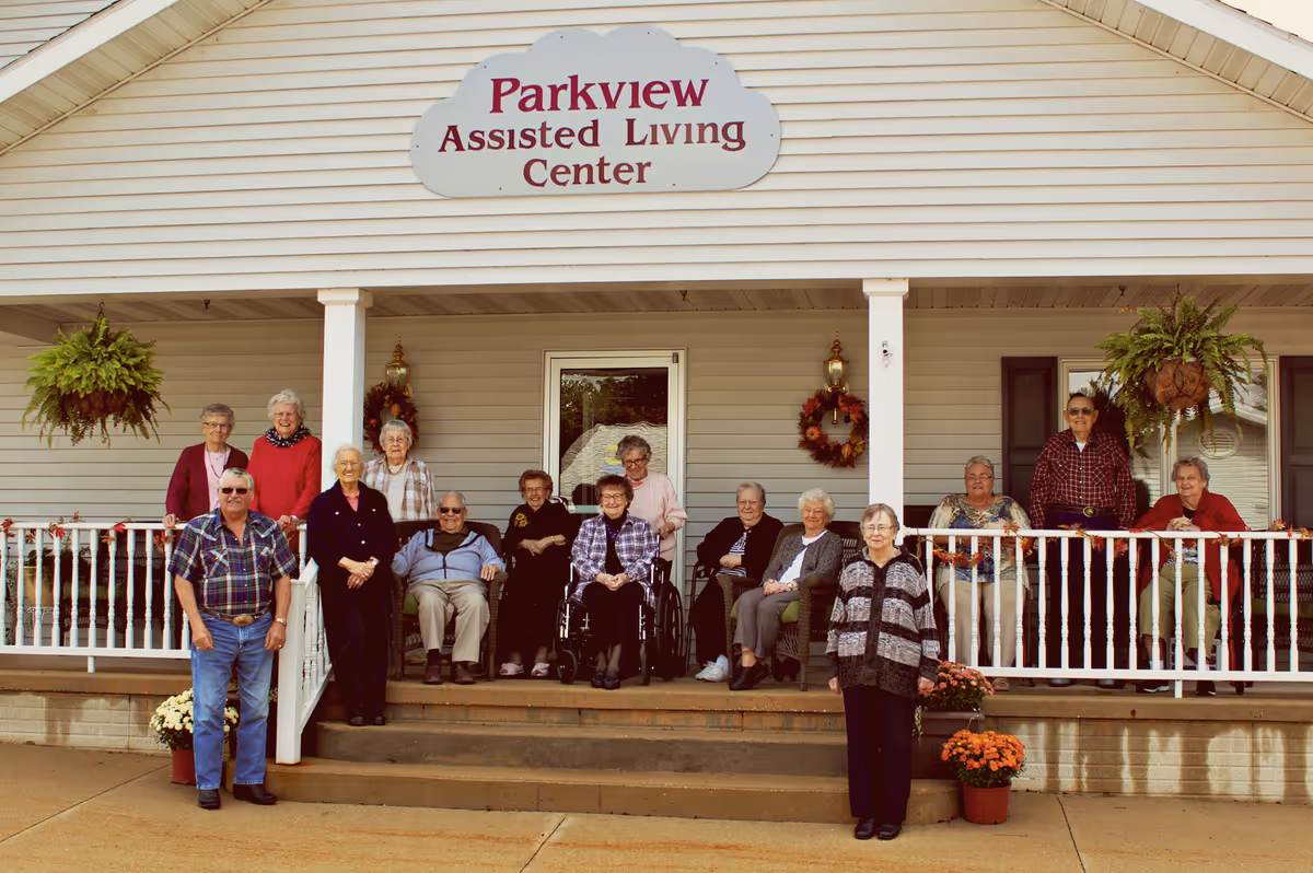 A group of elderly people posing together on the front porch of Parkview Assisted Living Center. The porch has white railings, hanging plants, and autumn wreaths on the walls. The group includes men and women, some seated and some standing, smiling at the camera.