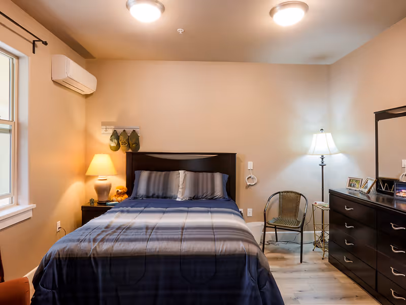 A cozy bedroom with a neatly made bed featuring a dark wooden headboard and blue-gray bedding. There is a nightstand with a lamp and a teddy bear on the left side of the bed. On the right side, there is a floor lamp, a wicker chair, and a dark wooden dresser with a mirror and framed photos. The room has beige walls, two ceiling lights, a window on the left, and a wall-mounted air conditioning unit.