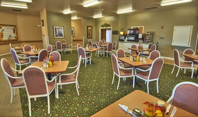 A dining room with multiple square tables, each surrounded by four cushioned chairs with pink upholstery and white frames. The room has a patterned green carpet and beige walls adorned with framed pictures. In the back, there is a beverage station with soda dispensers and a counter. The room is well-lit with ceiling lights.