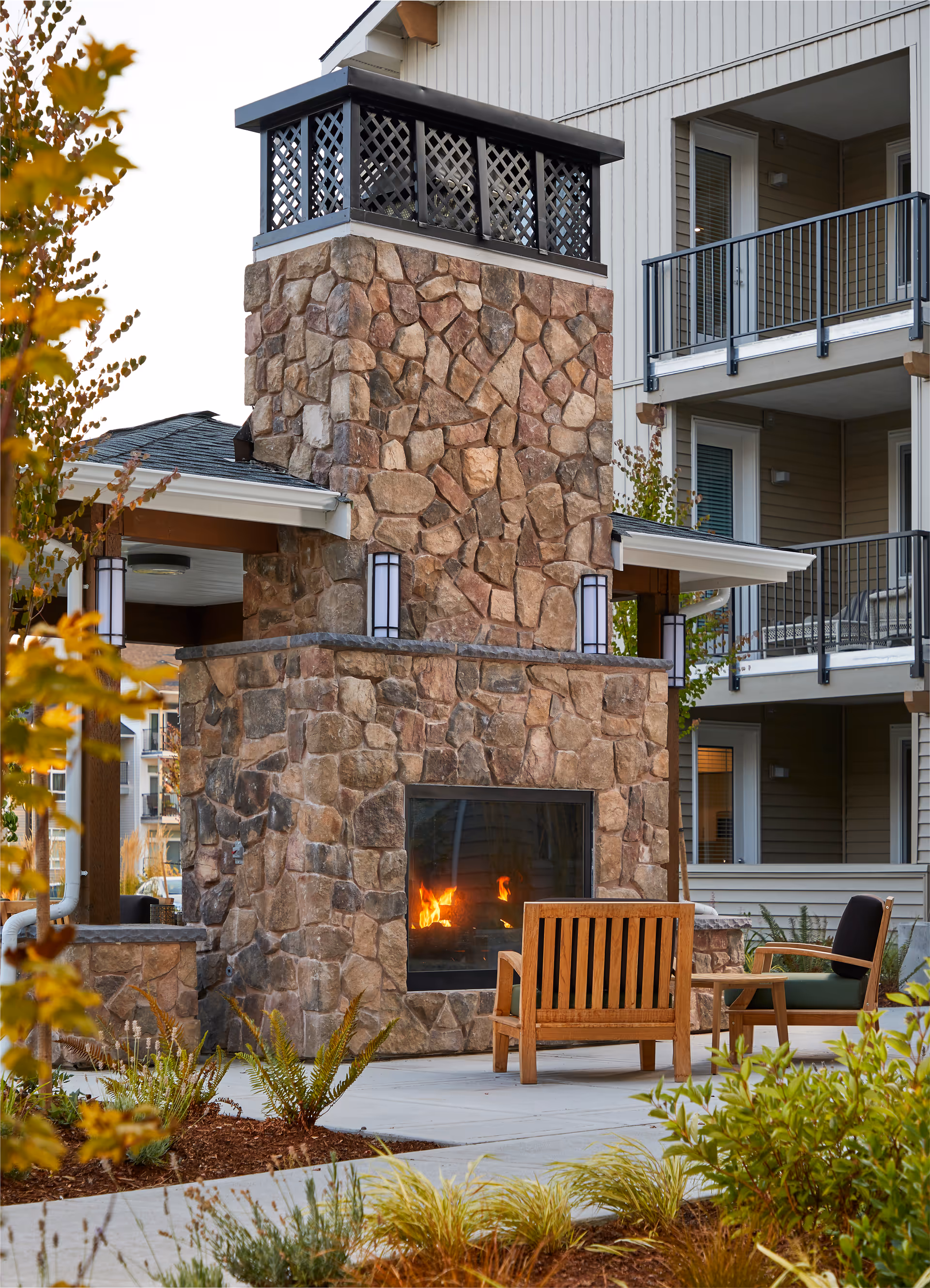 Outdoor seating area with a stone fireplace that has a fire burning inside. There are wooden chairs with cushions arranged around the fireplace. The area is surrounded by plants and is part of a residential building with balconies visible in the background.