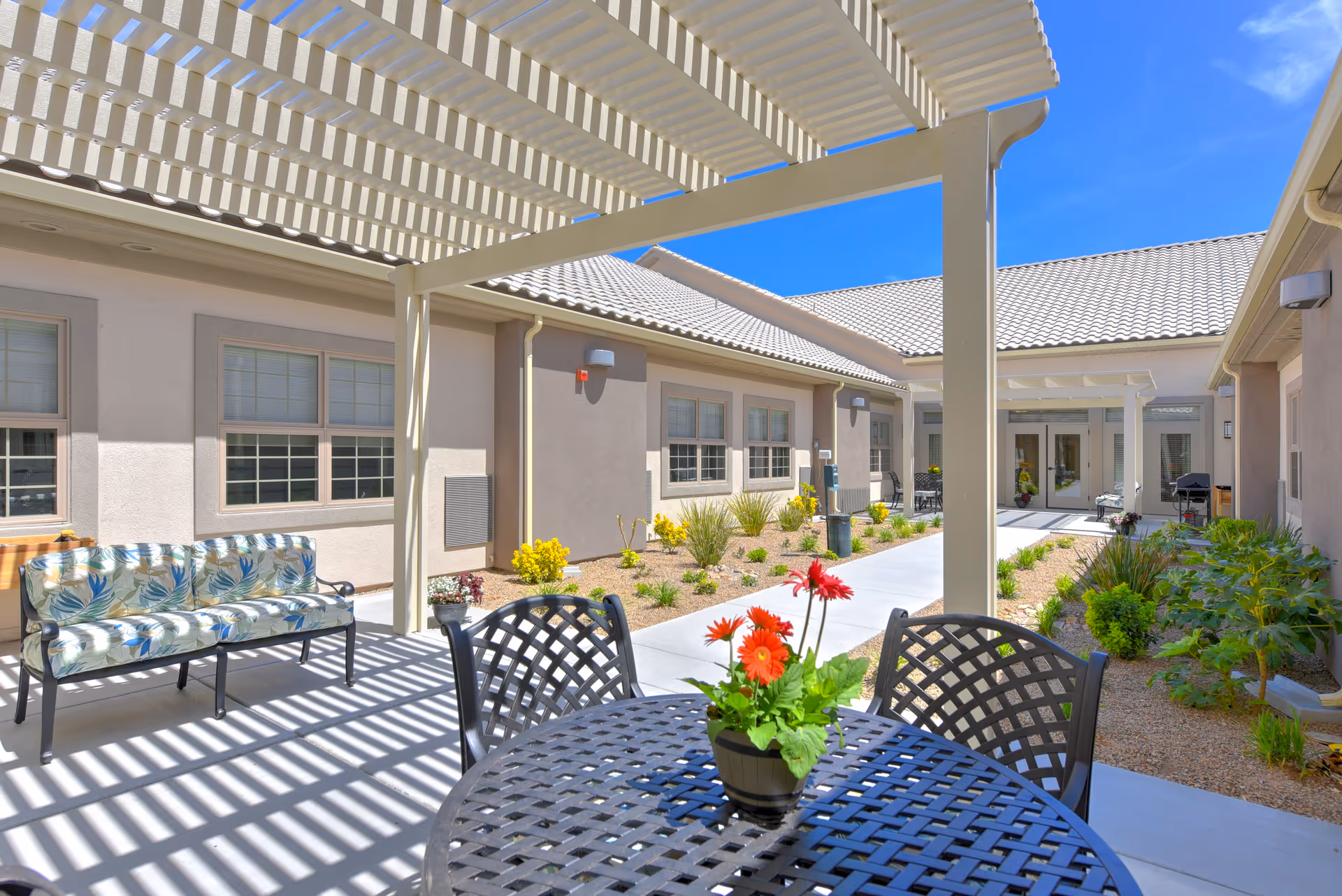 Outdoor patio area at Silverado Red Rock with a metal table and chairs, a potted plant with red flowers on the table, a cushioned bench with floral upholstery, and a pergola casting striped shadows. The patio is surrounded by beige buildings with tiled roofs and landscaped with small plants and shrubs under a clear blue sky.
