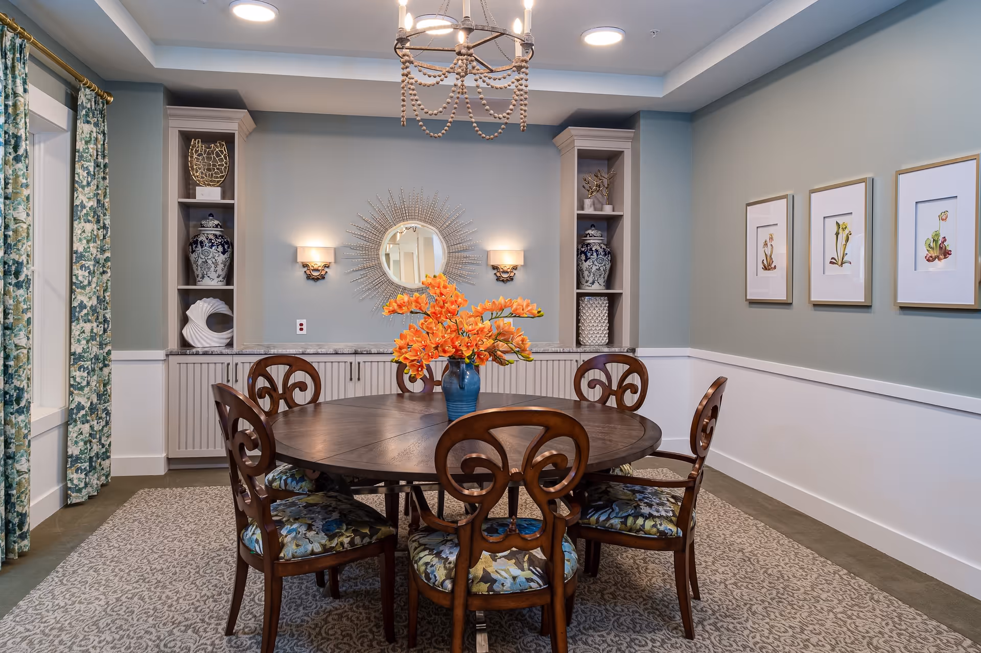 A dining room with a round wooden table surrounded by six ornate wooden chairs with floral upholstered seats. A blue vase with orange flowers is centered on the table. The walls are painted light blue with white wainscoting, decorated with three framed botanical prints on the right wall. Built-in shelves with decorative vases and sculptures flank a sunburst mirror with two wall sconces on the back wall. Floral patterned curtains hang on the left side, and a beaded chandelier hangs from the ceiling.