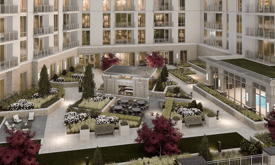 A landscaped outdoor courtyard area within a multi-story residential building. The courtyard features paved walkways, garden beds with white flowers and green shrubs, benches, a seating area with tables and chairs under a pergola, and several trees with red foliage. The building surrounding the courtyard has balconies and large windows, and there is a glass-enclosed room with a green roof on the right side.