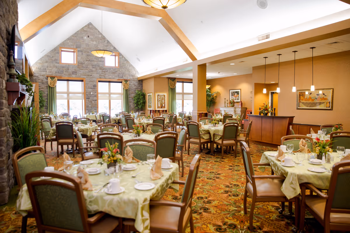A spacious and well-lit dining room with multiple round tables covered in light green tablecloths, each set with plates, cups, glasses, and napkins. The room features high vaulted ceilings with wooden beams, large windows with green curtains, a stone accent wall, and warm lighting fixtures. The carpet has a colorful floral pattern, and there are plants and framed artwork decorating the space.