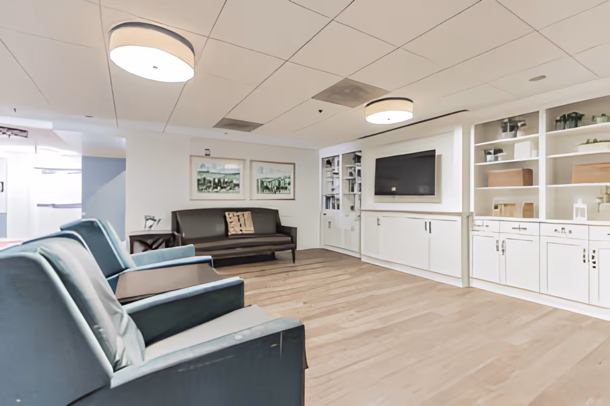 A bright and spacious living room area with light wood flooring, two blue armchairs in the foreground, a dark brown sofa against the far wall, and built-in white cabinetry with shelves and a large flat-screen TV mounted on the wall. The ceiling has recessed lighting and two round ceiling lights.