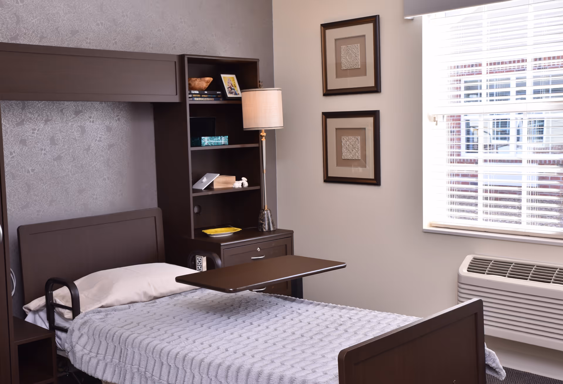 A single bed with a gray blanket and white pillow in a room. Next to the bed is a dark wooden nightstand with a lamp, shelves holding books, a framed photo, and decorative items. Two framed artworks hang on the wall near a window with white blinds. Below the window is an air conditioning unit.