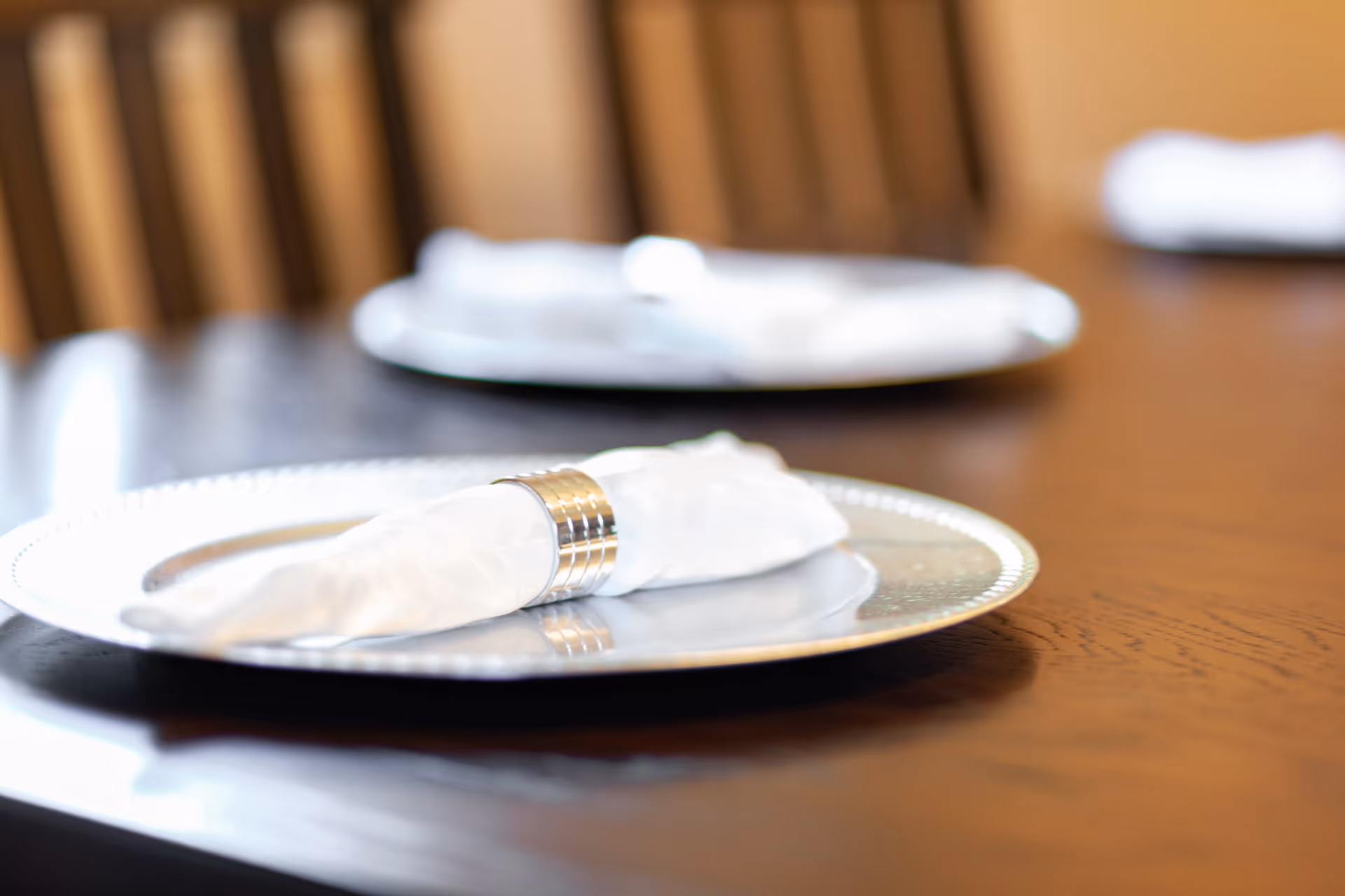 Close-up of a dining table set with a silver charger plate and a white cloth napkin held by a silver napkin ring, with blurred plates and napkins in the background.