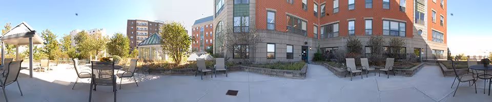 Outdoor patio area at Assisted Living Residences at Hancock Park with several tables and chairs arranged on a concrete surface. The patio is surrounded by a multi-story brick building and landscaped with small trees and shrubs under a clear blue sky.