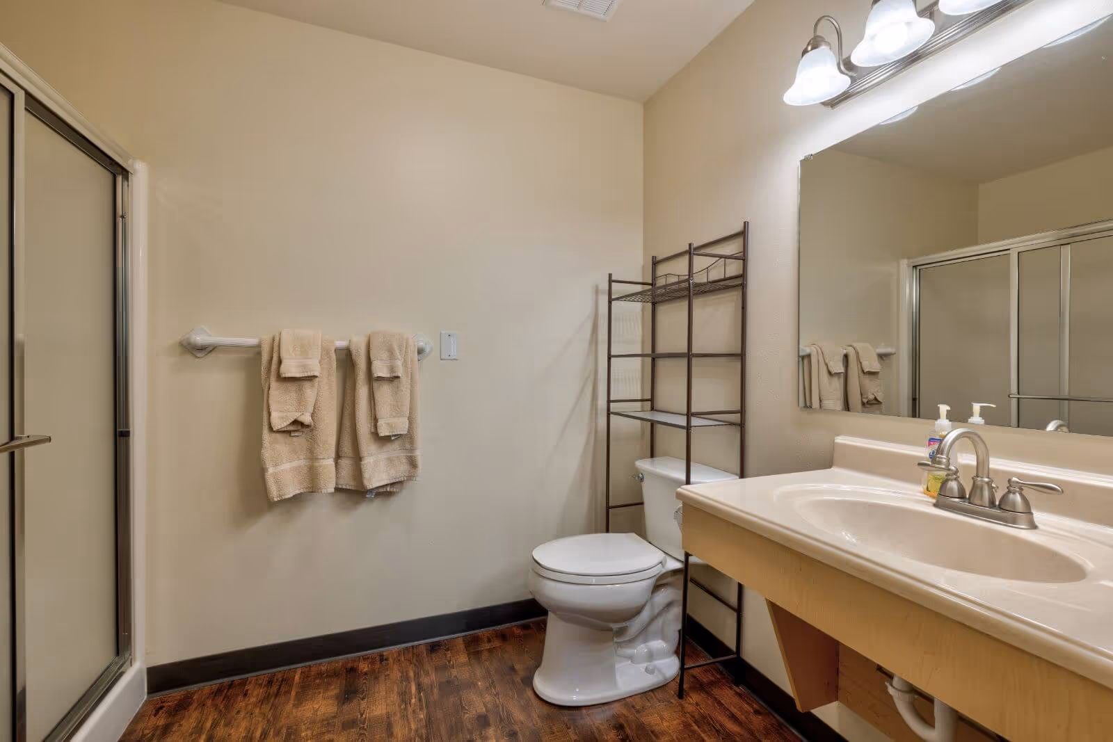Bathroom with a sink vanity and mirror, toilet with a metal shelving unit above it, glass-enclosed shower, and a towel rack holding beige towels.