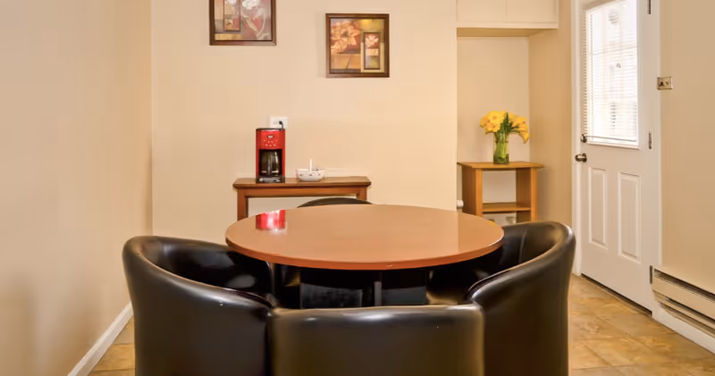 A small dining area with a round wooden table surrounded by four black leather chairs. In the background, there is a small wooden table with a red coffee maker and a bowl on it, and a corner table with a vase of yellow flowers. Two framed floral paintings hang on the beige wall, and a white door with a window is visible on the right side.