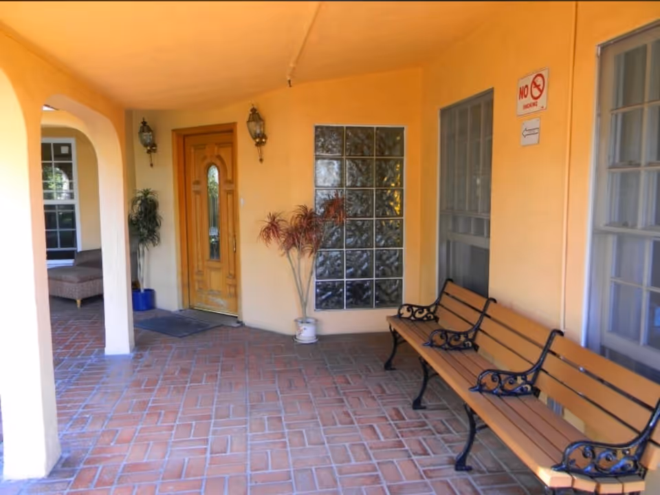 Covered outdoor patio area with a wooden door, two wall-mounted lanterns, potted plants, a glass block window, a no smoking sign, and a wooden bench with black metal armrests and legs on a tiled floor.