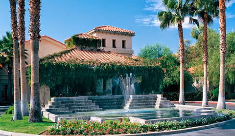 Exterior view of a retirement community building with a terracotta tiled roof, surrounded by palm trees and greenery. In front of the building is a large tiered water fountain with water jets, bordered by a well-maintained garden with flowers and shrubs under a clear blue sky.