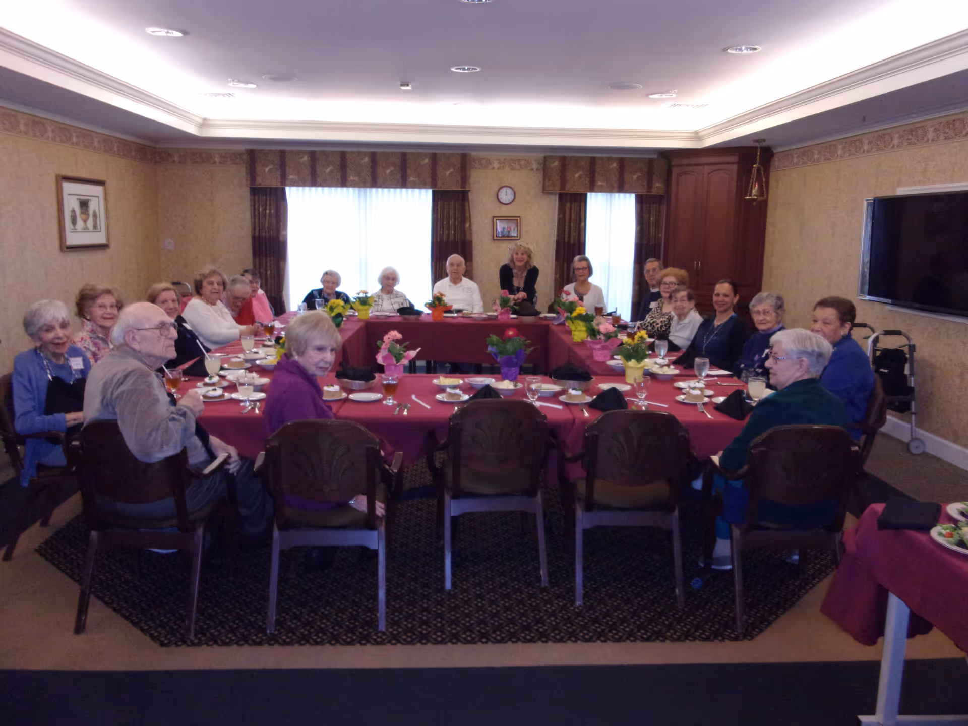 A group of elderly people seated around a large U-shaped table in a dining room. The table is covered with a red tablecloth and decorated with colorful potted flowers. Plates, glasses, and utensils are set on the table. The room has beige wallpaper, a clock, framed pictures on the wall, and a large window with curtains letting in natural light.