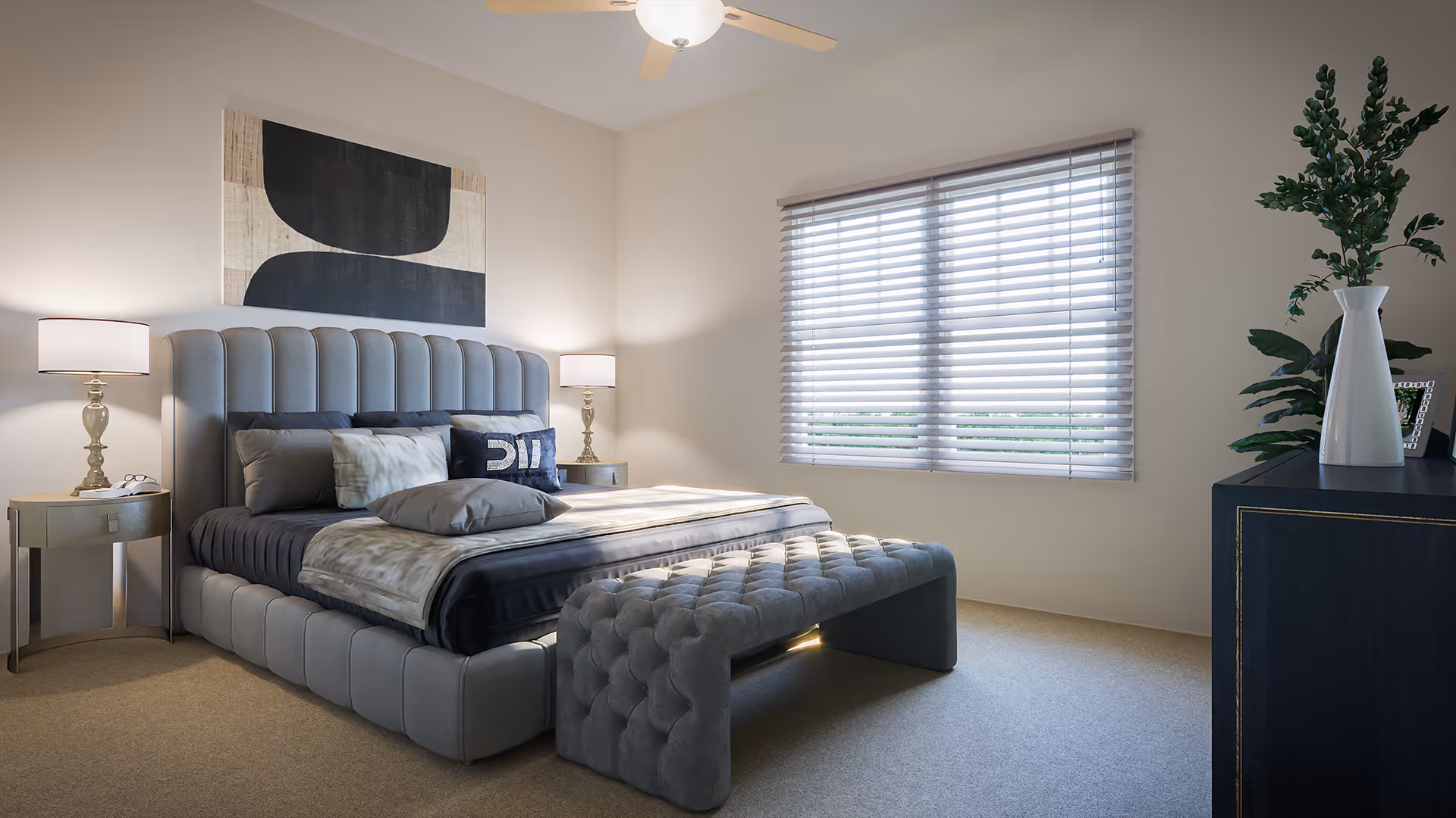 A modern bedroom with a large upholstered bed featuring multiple pillows and a gray quilted bench at the foot. Two bedside tables with matching lamps flank the bed. A large window with horizontal blinds allows natural light to fill the room. A dark dresser with a white vase and green plant is positioned against the right wall. The walls are painted light beige, and a ceiling fan with a light fixture is mounted above.