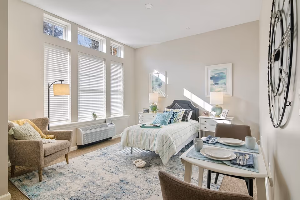 A bright and cozy bedroom with a single bed covered in light striped bedding and multiple pillows. The room features a large window with white blinds, a beige armchair with a pillow and throw blanket, two white nightstands with lamps and framed pictures, a small dining table set for two with plates, cups, and silverware, and a large decorative wall clock. The floor is covered with a patterned area rug.